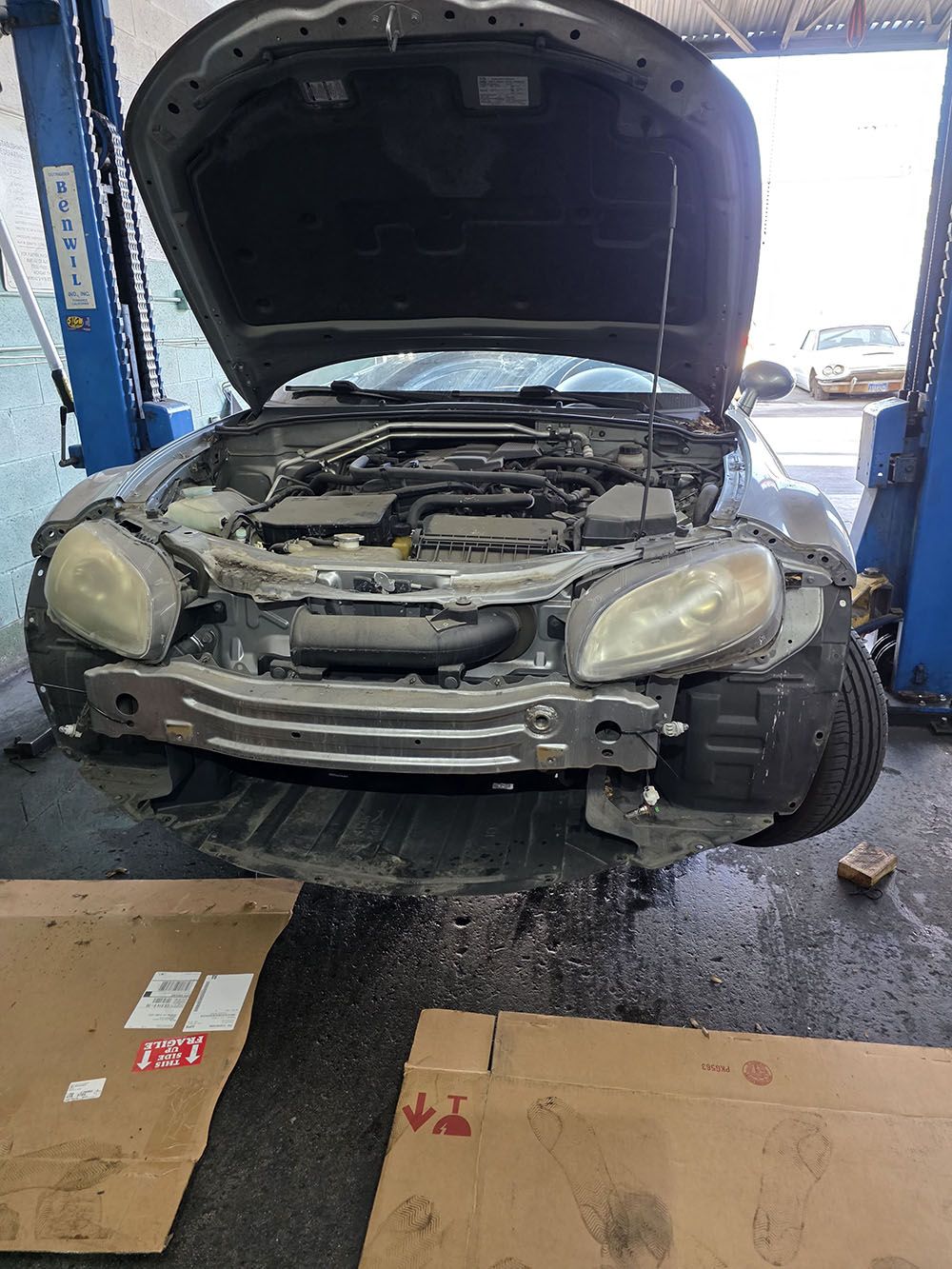 Damaged gray car with hood open in an auto repair shop, front bumper removed and parts exposed
