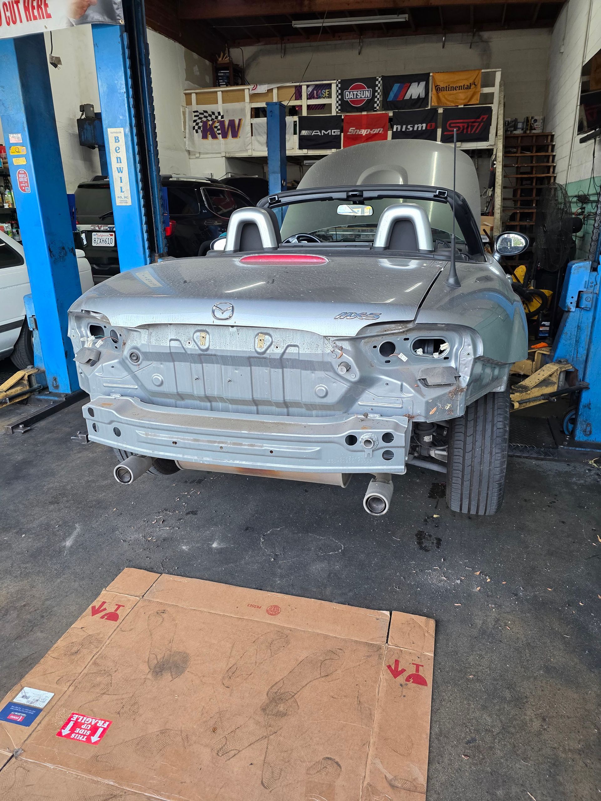 Gray race car body in an auto shop, front bumper removed and car on a lift with tools nearby.