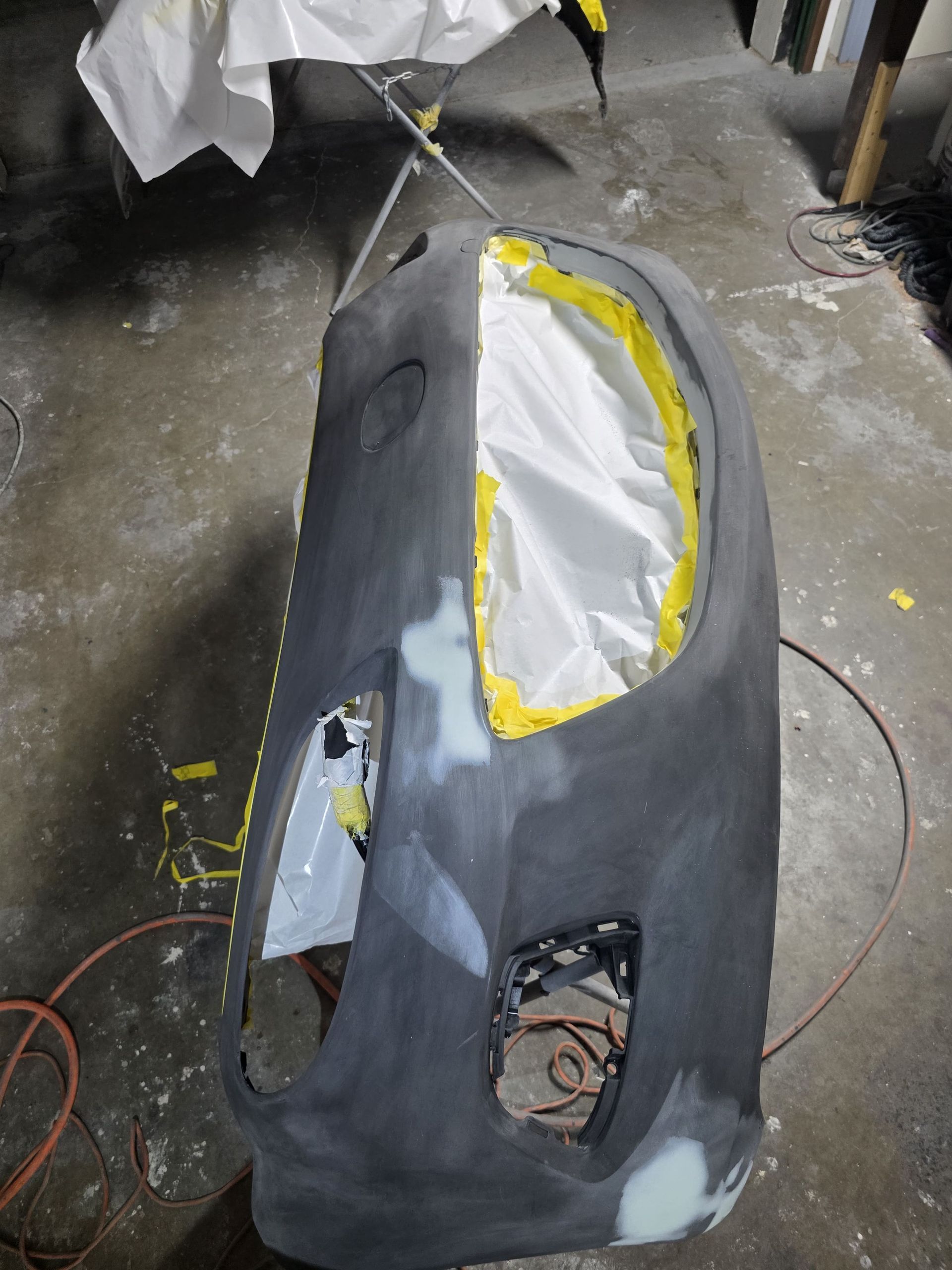 Black car bumper/fender panel with yellow tape and white filler in a workshop, showing sanded repair areas