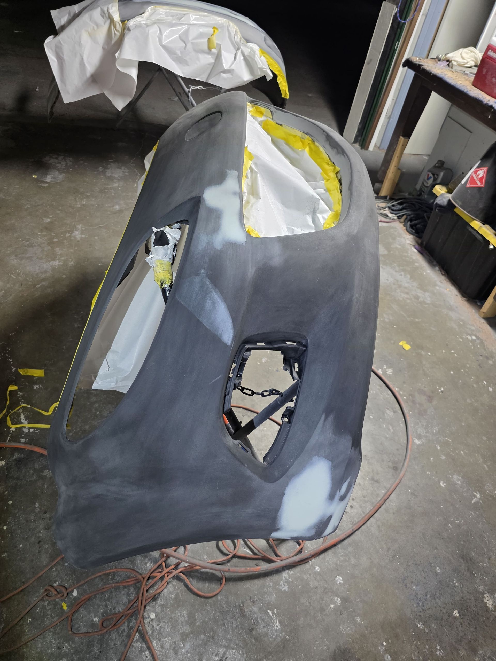 Gray car body panel in a workshop, with taped edges and cutout openings for windows and lights.