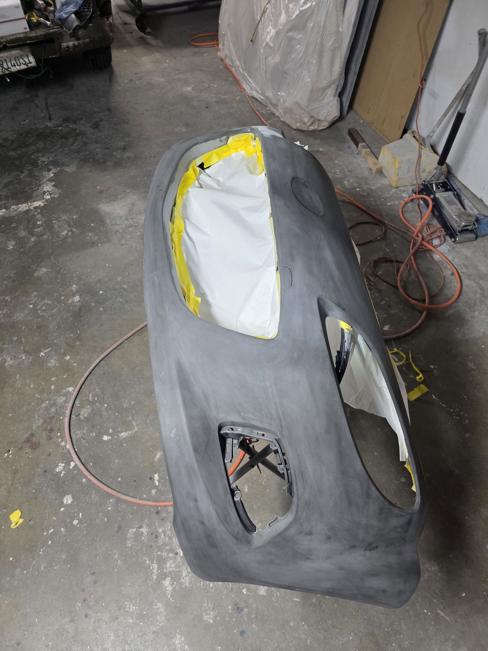Gray car bumper primed for repainting, with masking tape and cutout openings in a workshop.