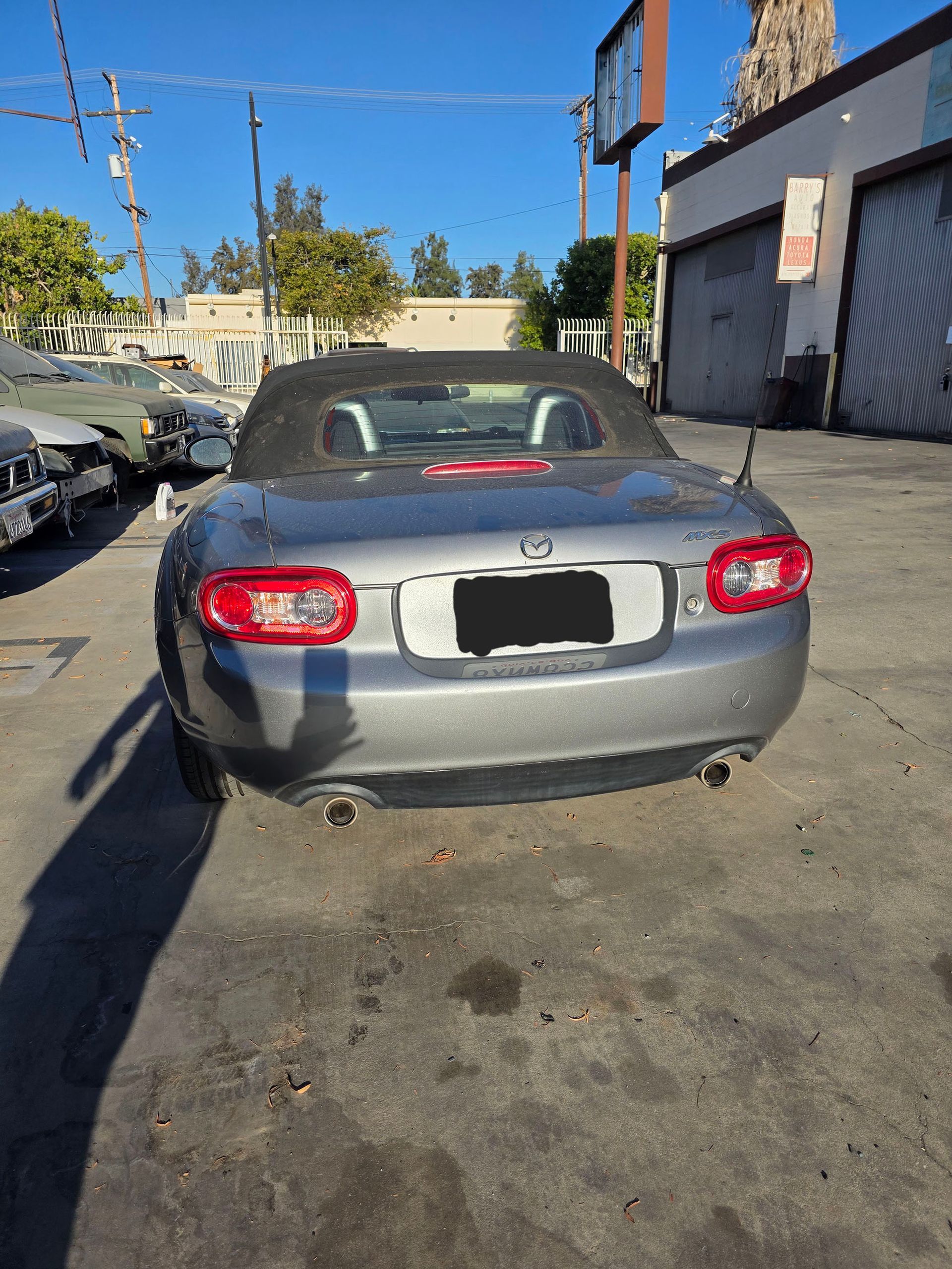 Rear view of a gray convertible sports car parked in an outdoor lot, with damage on the left rear bumper.