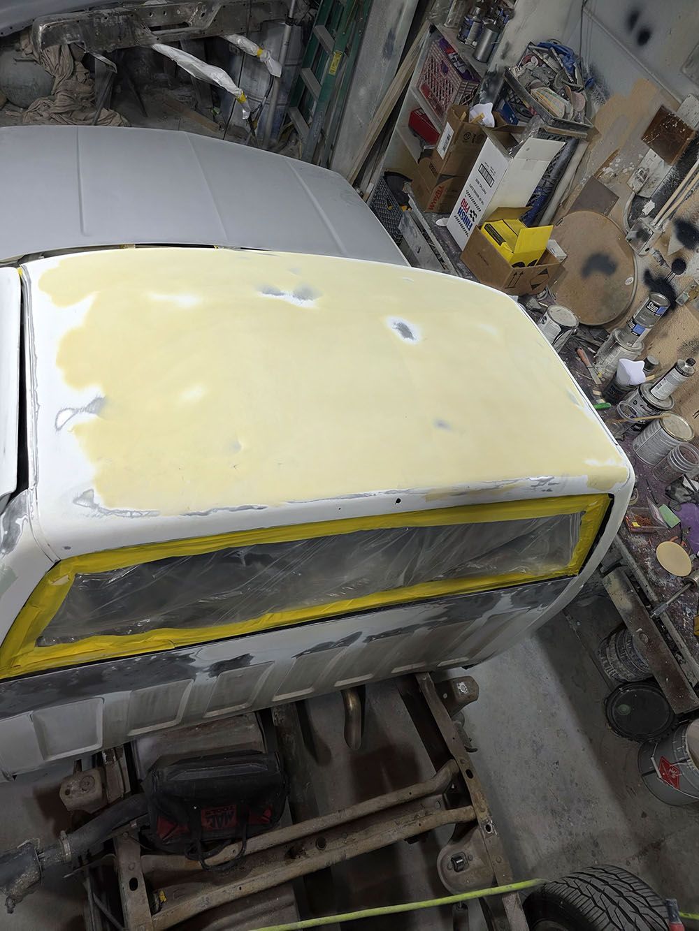 Car body in auto shop being sanded and primed, with yellow-taped windows and a light gray roof.