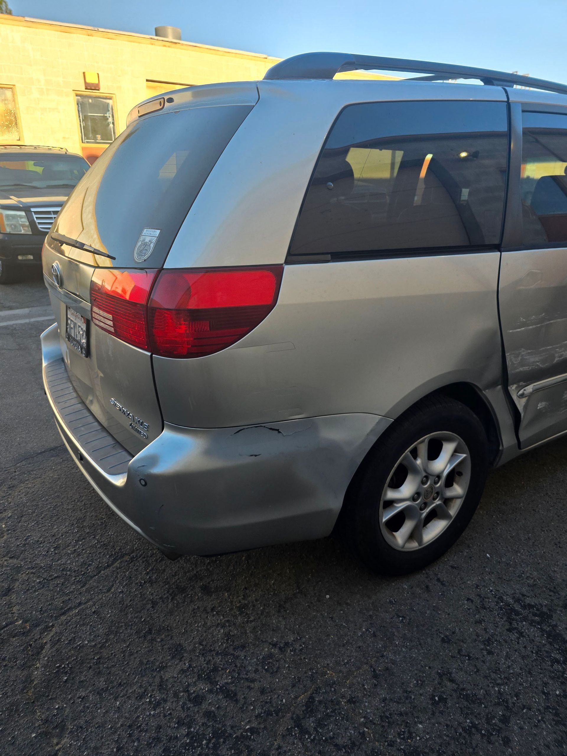 Gray minivan parked in a lot, rear side view, with a red taillight and sunset light reflecting off it