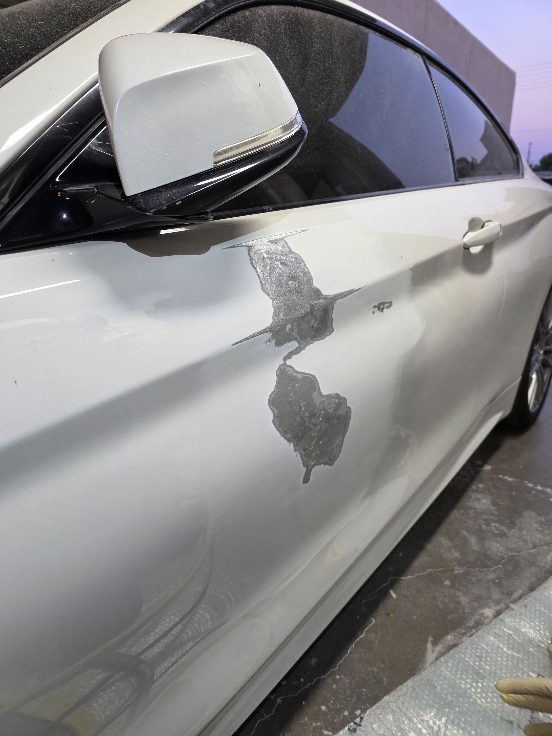 White car door with a large gray paint scrape and damage near the side mirror.