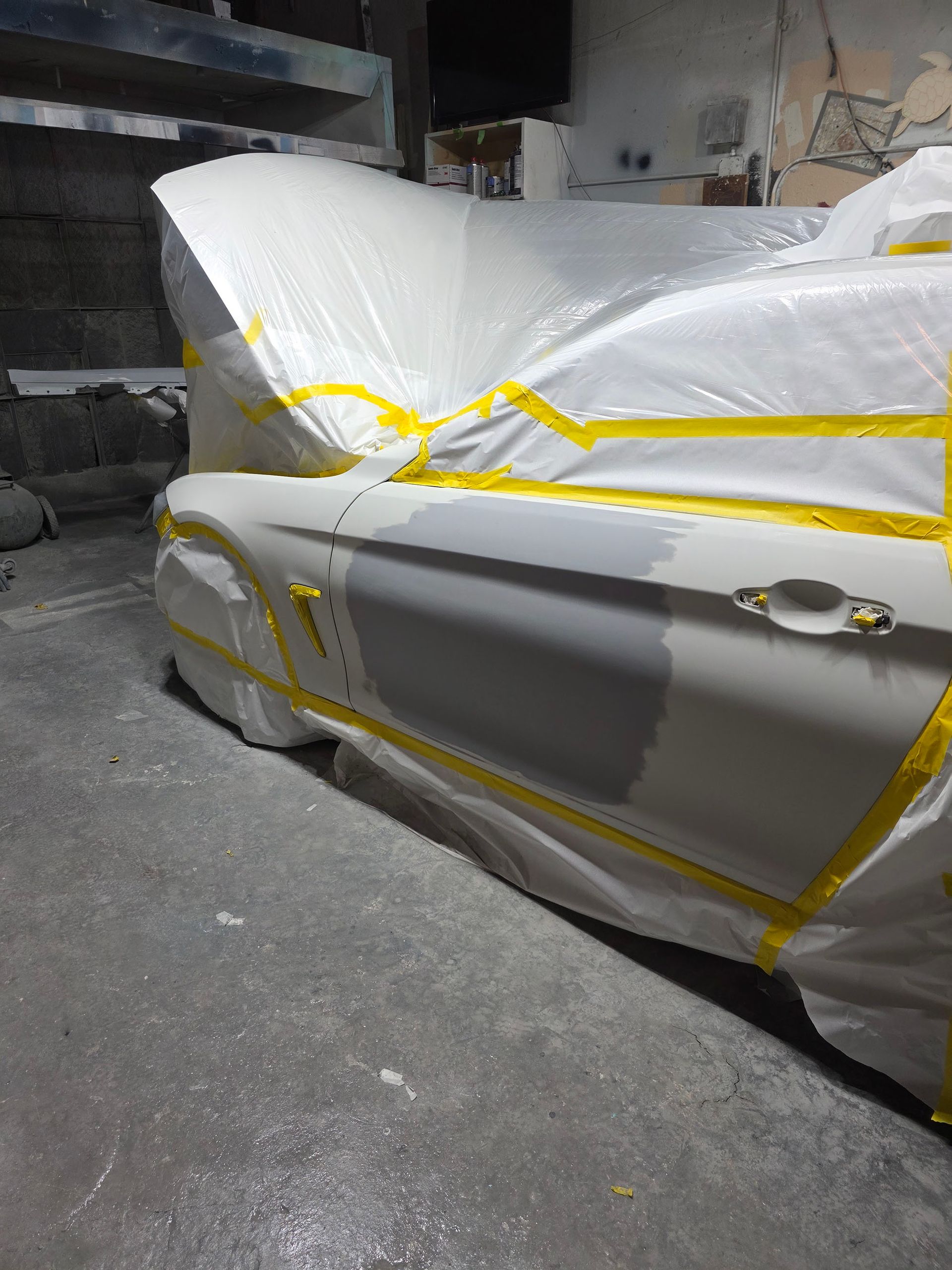 Car covered in white plastic and yellow tape, parked in a dim garage.