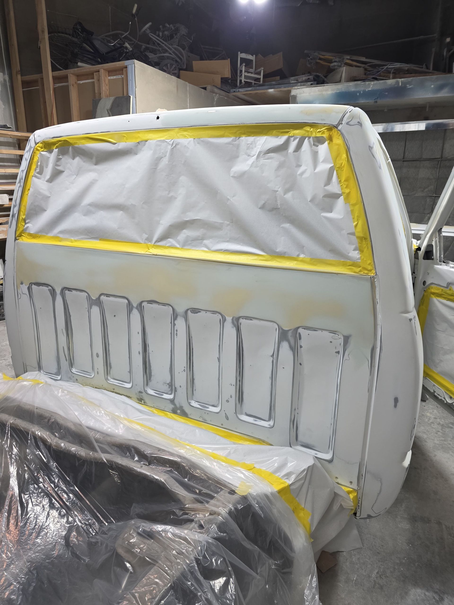 White car body part indoors, wrapped in plastic and masked with yellow tape for painting