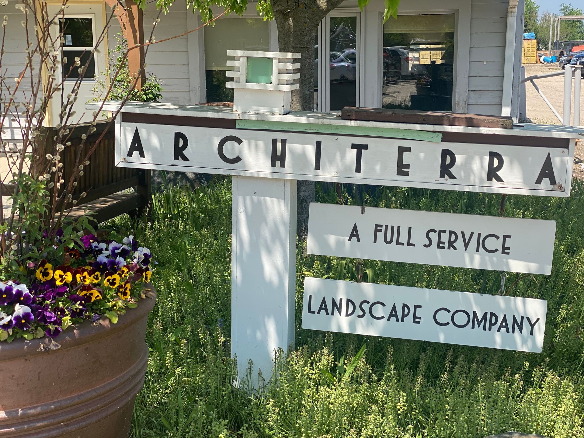 Sign for Architerra Landscape Company. White sign with text, flowers in a pot, and a building in the background.