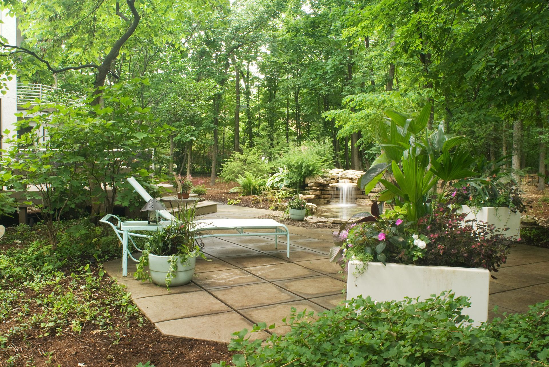 Patio with lounge chairs, planters, and small waterfall surrounded by lush greenery and trees.