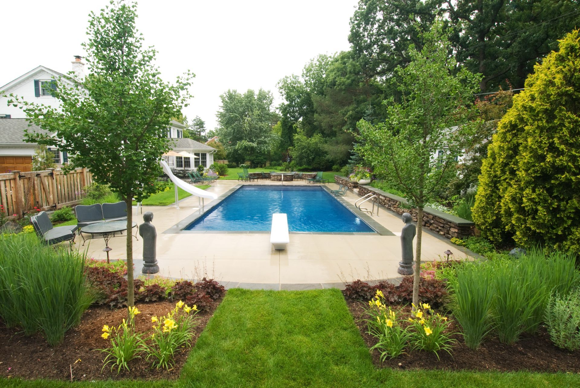 Backyard with a rectangular pool, lawn, and surrounding landscaping. Trees frame the pool area.