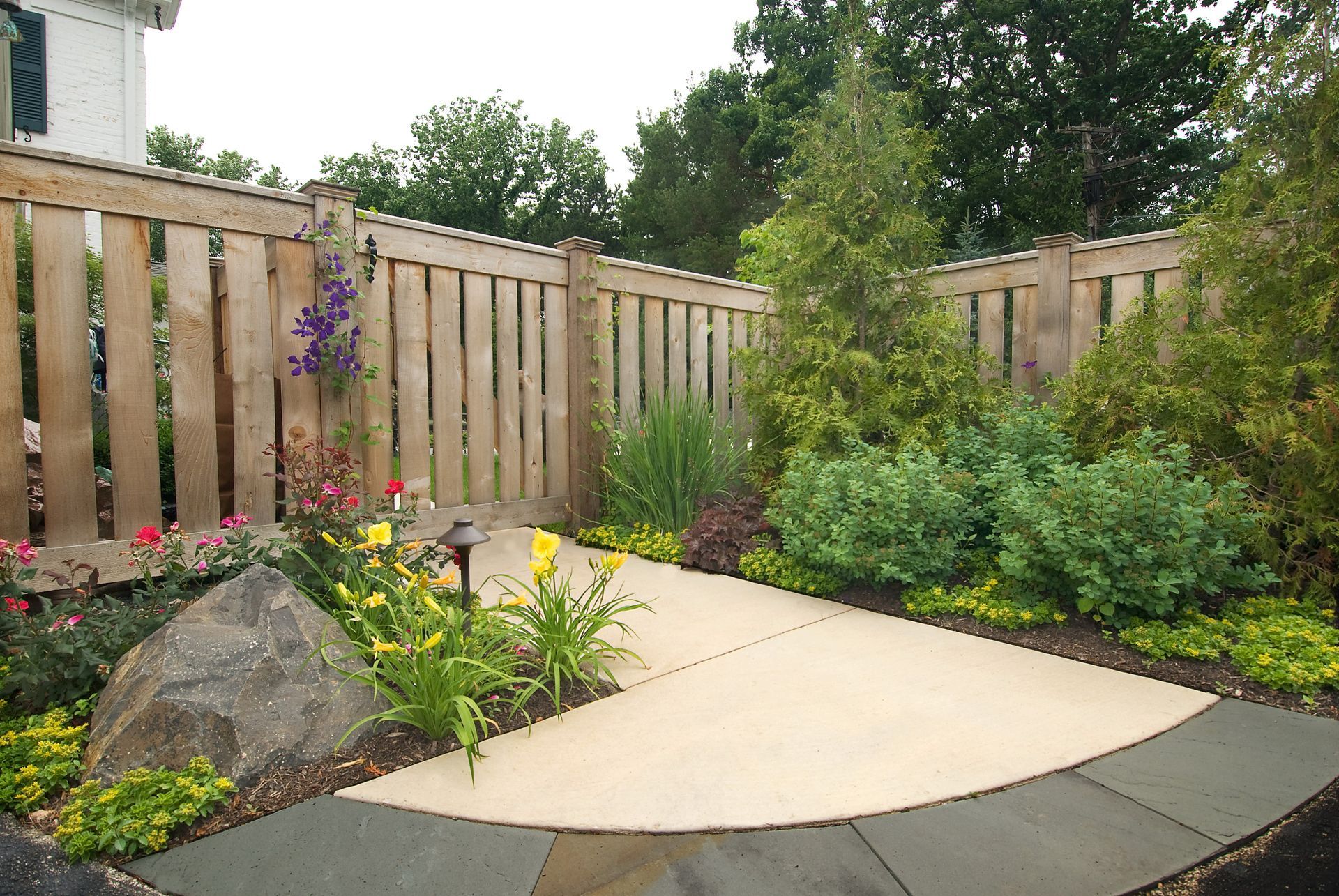 A garden path framed by a wooden fence, with flowers, shrubs, and a large rock.