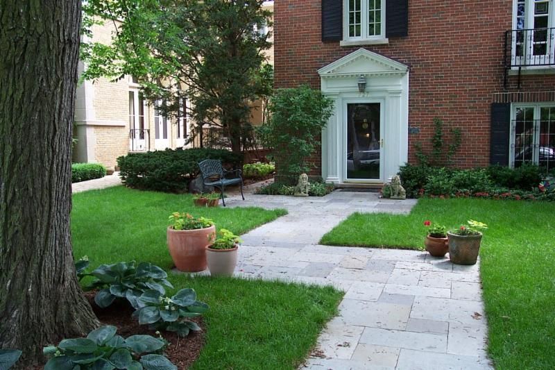 Lush green lawn with a stone path leading to a brick building's entrance. Potted plants and a tree.