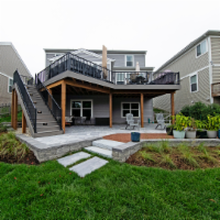 Two-story deck with stairs, overlooking a patio and backyard. House is light gray with black railing.