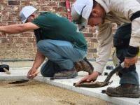 Two workers installing pavers outdoors. One squats, the other kneels, both using tools.