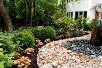 Stone pathway curves through a garden with shrubs, plants, and a white building.