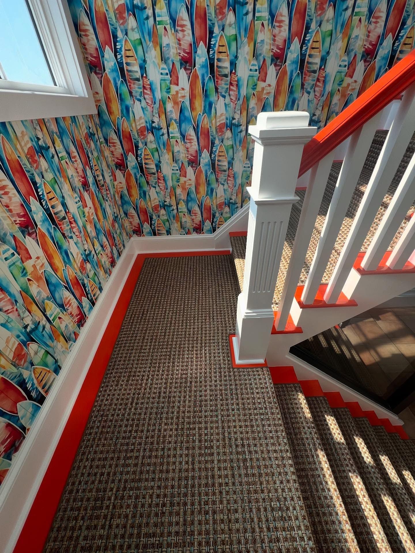 Staircase with colorful wallpaper and red trim. Brown woven carpet and white railing with red accents.