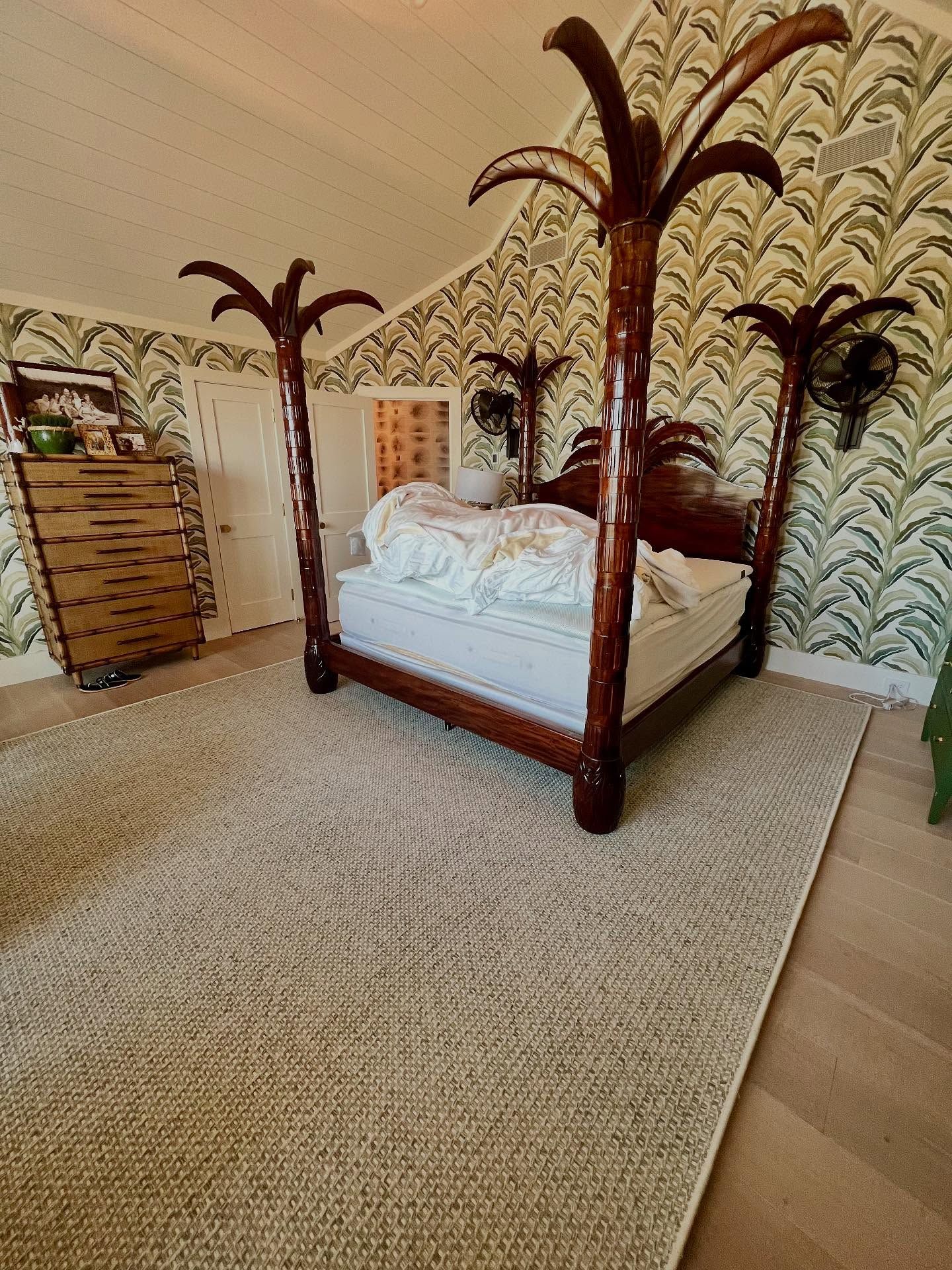 Bedroom with a four-poster bed shaped like palm trees, a dresser, and patterned wallpaper. Beige rug on the floor.