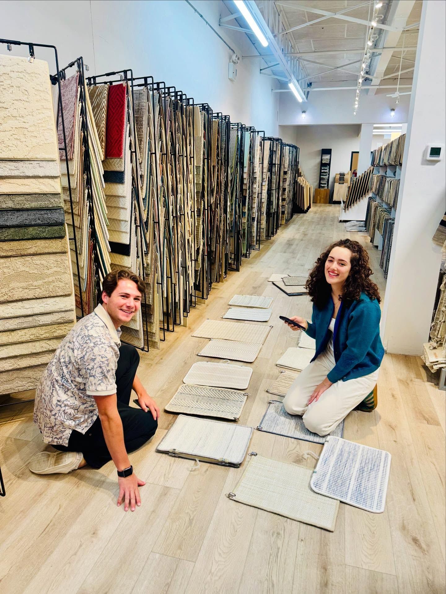 Two people kneel by carpet samples in a store, smiling. Carpet samples line shelves in the background.