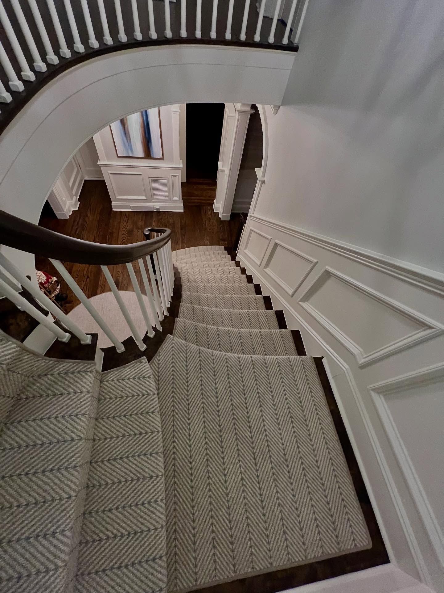 Staircase with herringbone patterned carpet, white walls, and dark wood handrail, viewed from above.
