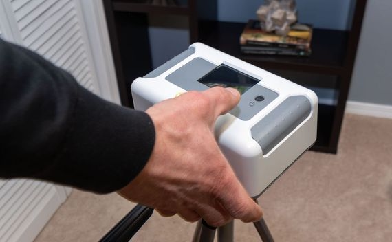 A person's hand holding a white and gray device on a tripod in a room. The device appears to have a screen.