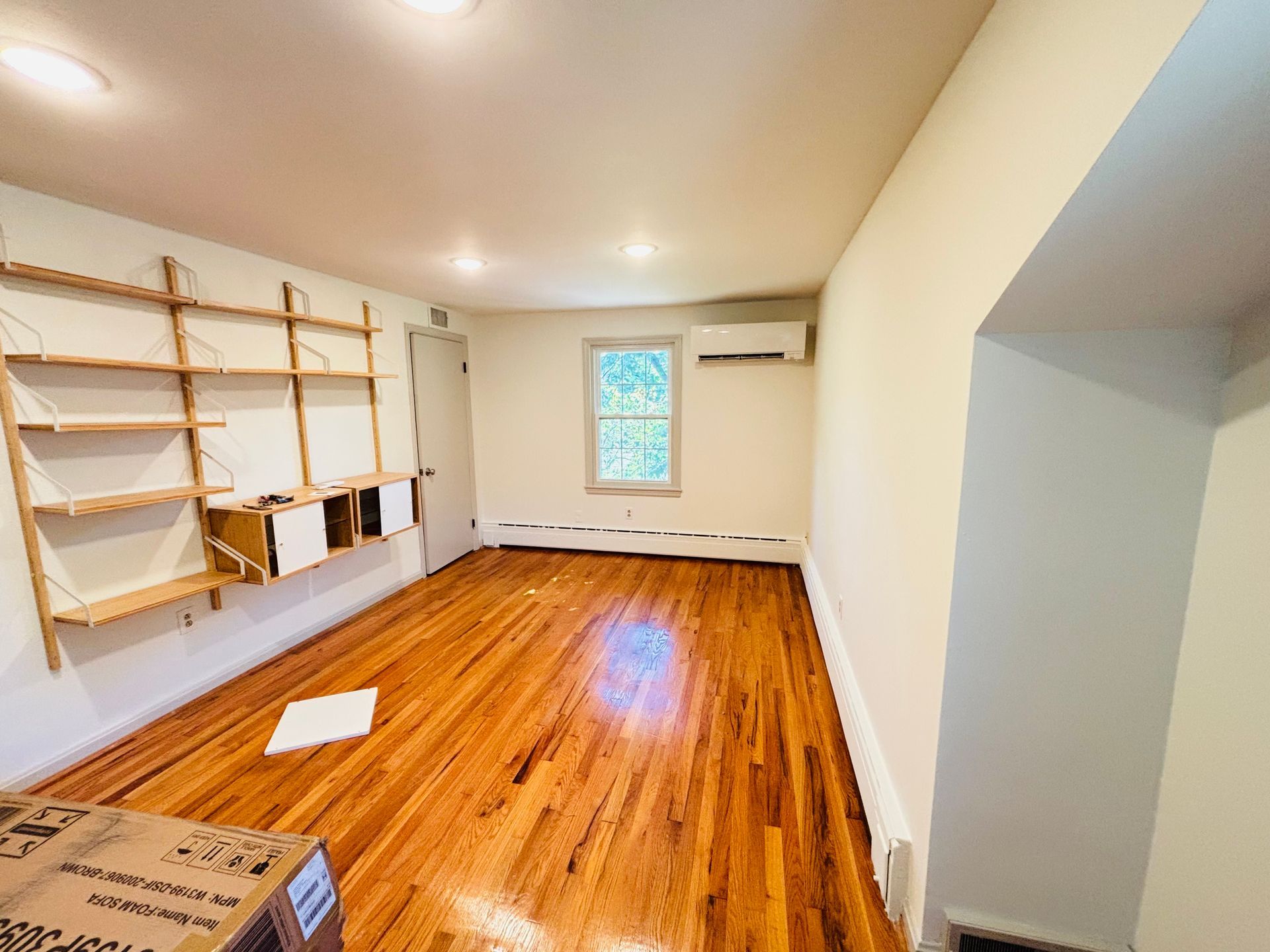 Empty room with wood floors, shelves on the wall, and a window.