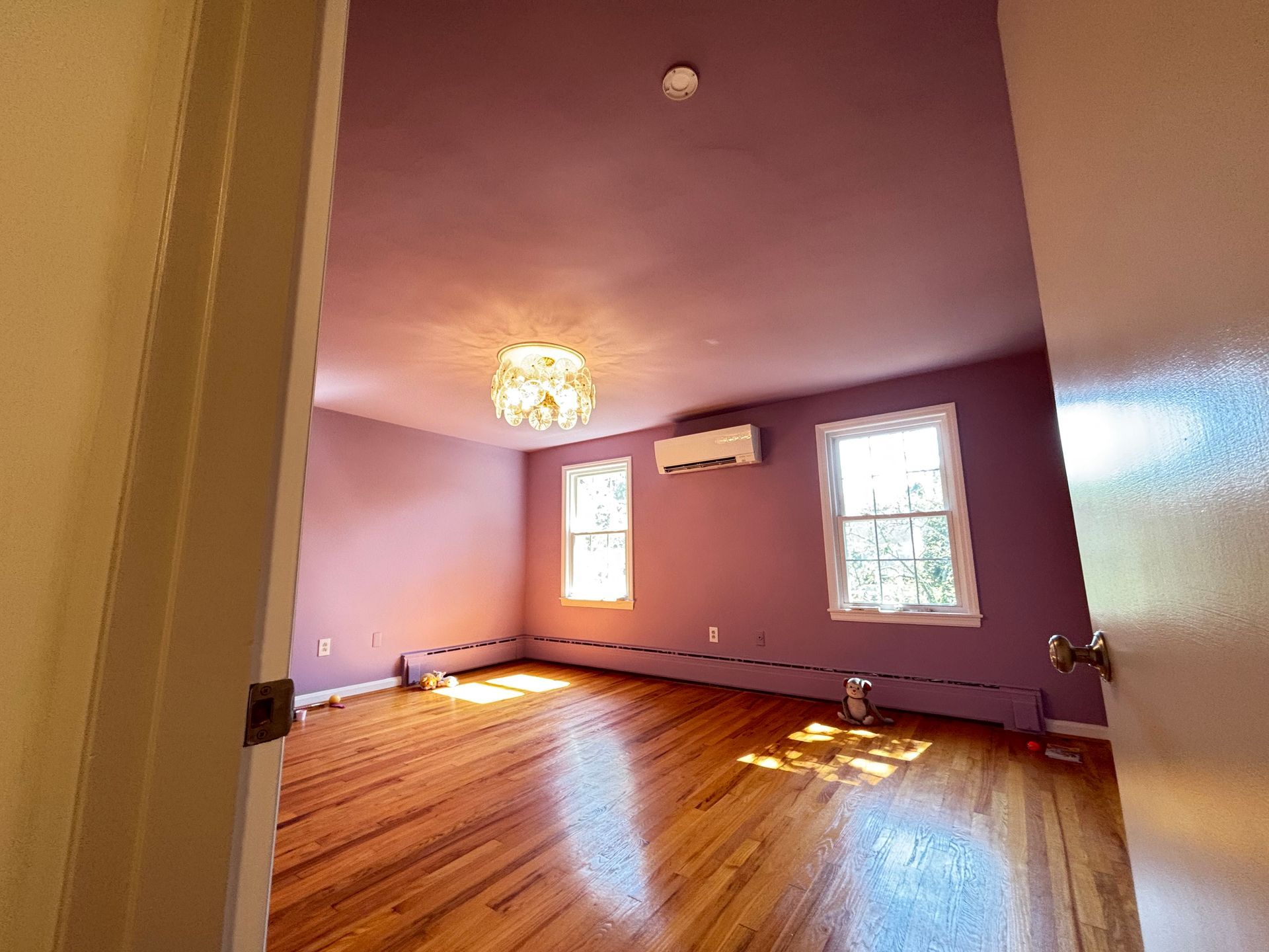 A room with purple walls, two windows, wood floor, chandelier, and AC unit.