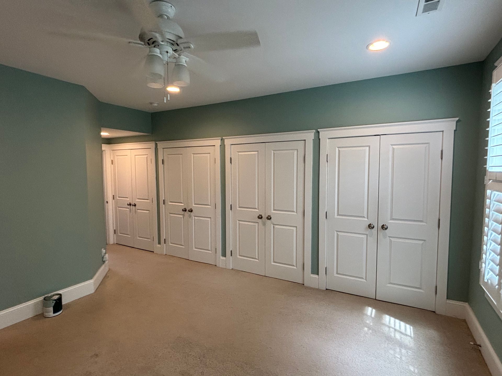 A room with white closet doors and a light green wall. Beige carpet, ceiling fan, and window with shutters.