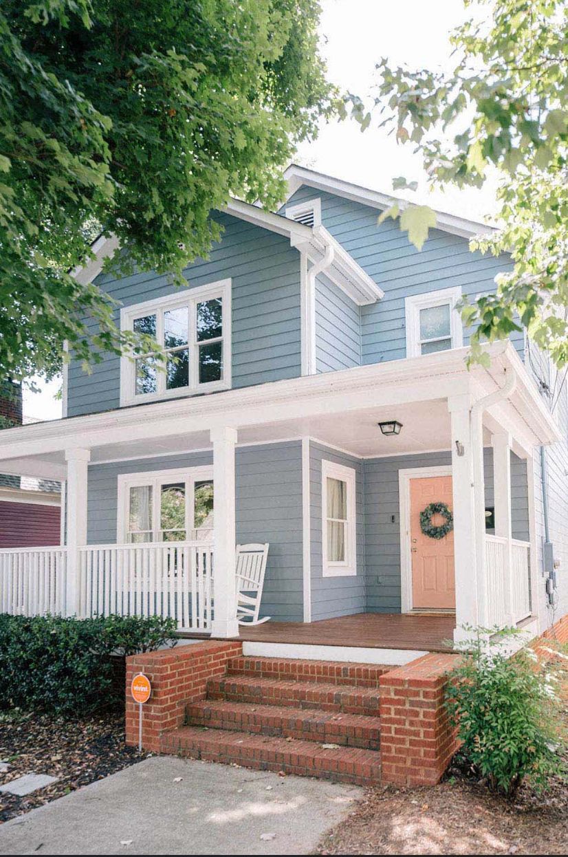 Two-story blue house with white trim, porch, pink door, and brick steps; trees frame the view.
