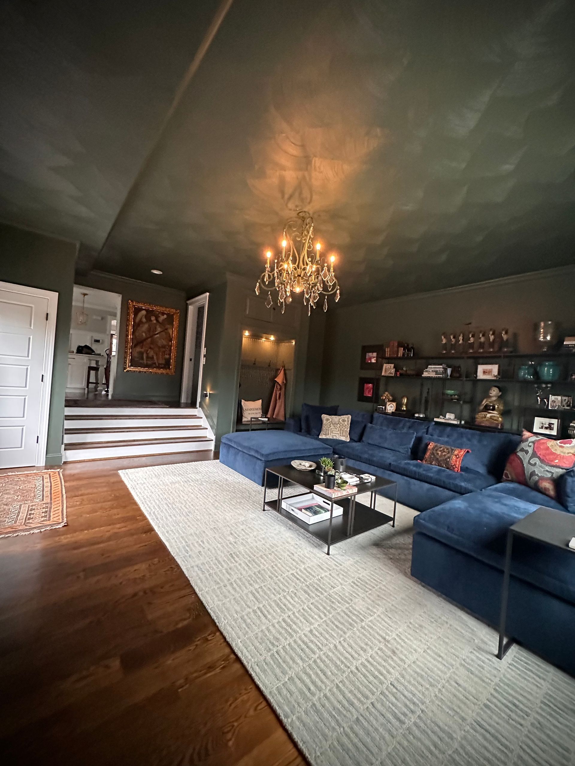 Cozy living room with dark green walls, blue sectional sofa, chandelier, and a white rug.