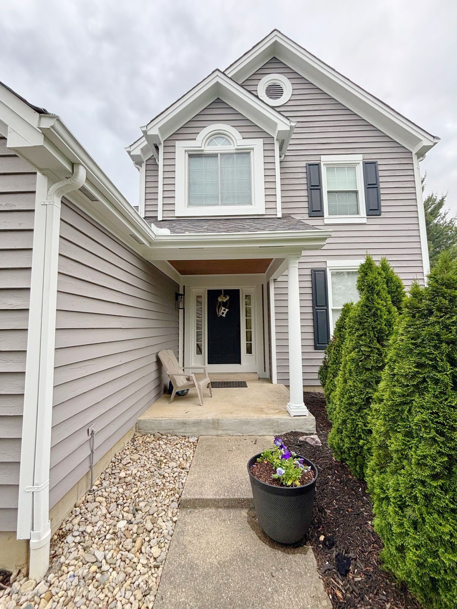 Two-story gray house with white trim, porch, and a stone walkway. Green bushes line the right side of the house.