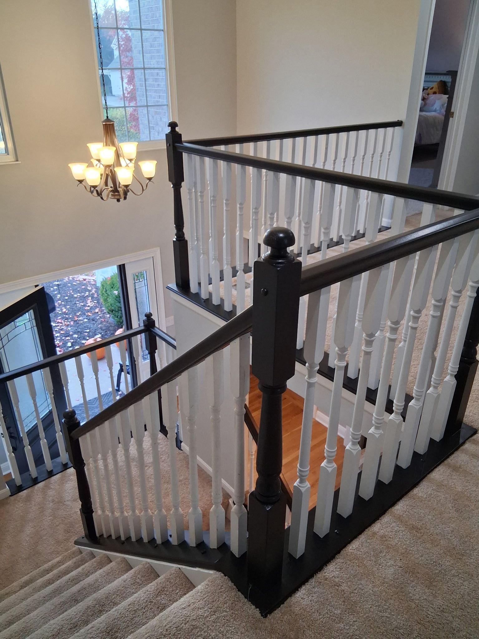 Staircase with dark-painted newel posts and handrails, white spindles. Beige carpet covers the steps. A chandelier hangs above.