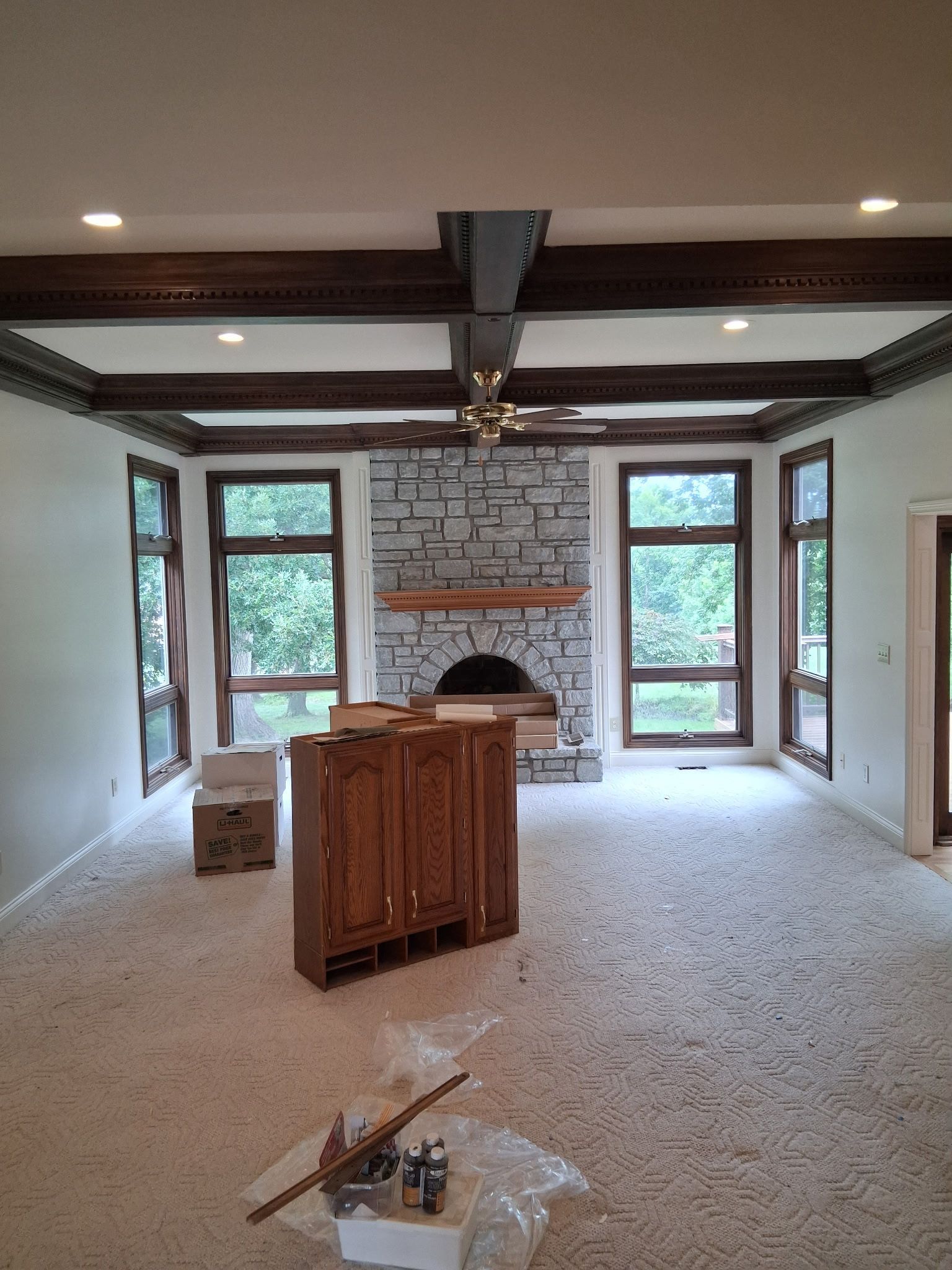 Spacious living room with stone fireplace, wood beams, windows, and light-colored carpet. A wooden cabinet stands in the center.