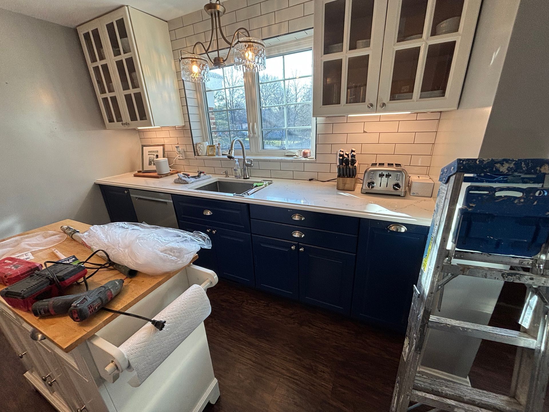 Kitchen with blue cabinets, white countertops, subway tile backsplash, and a window. A work island and ladder are present.