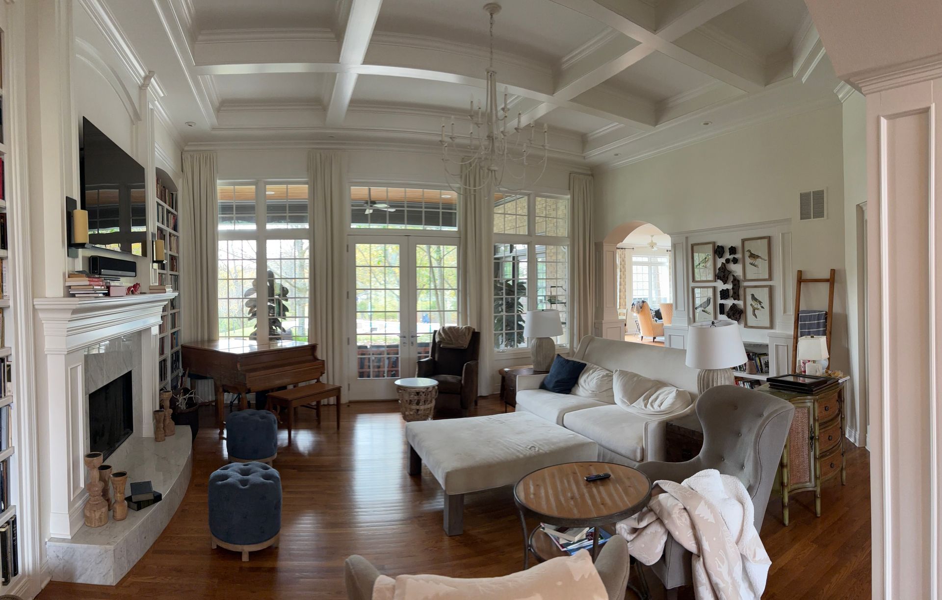 Spacious living room with white walls, fireplace, piano, windows, and chandelier. Wooden floors, a white couch, and various decor.