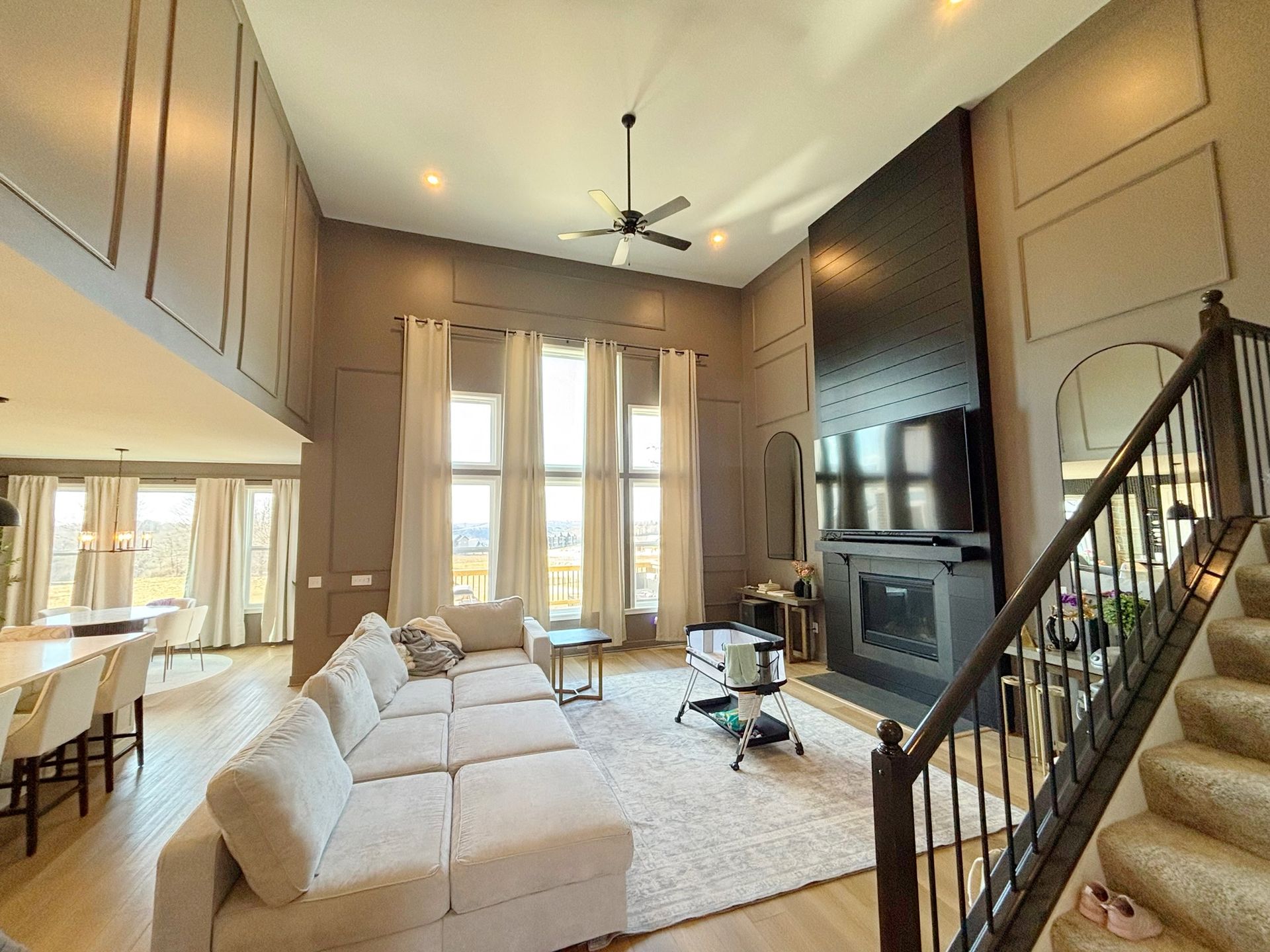 Spacious living room with high ceilings, large windows, and a black fireplace with TV, staircase on the right.