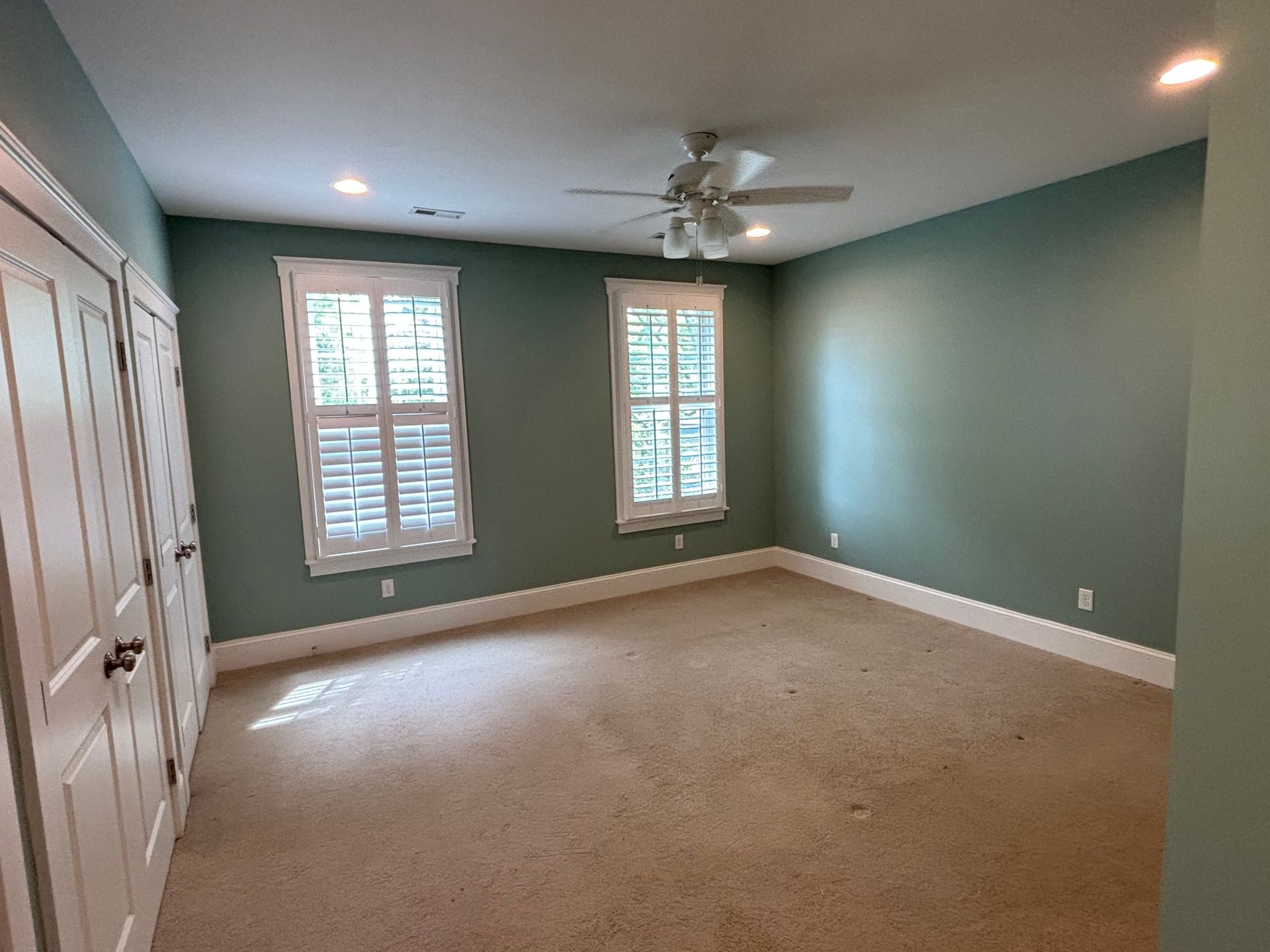 Empty bedroom with green walls, two windows with shutters, and tan carpet.