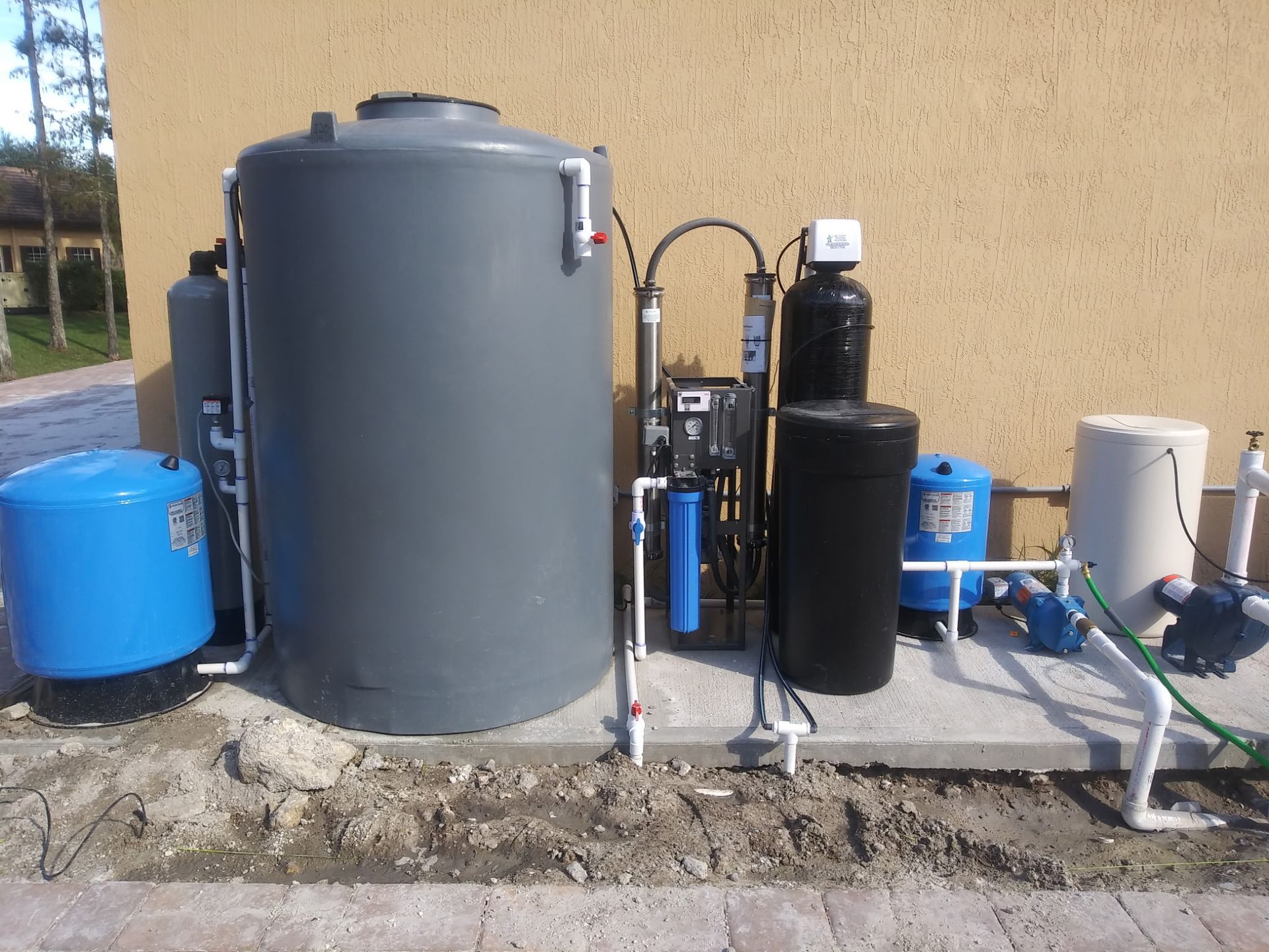 Outdoor water treatment equipment with gray and blue tanks, pipes, and pumps beside a beige wall.