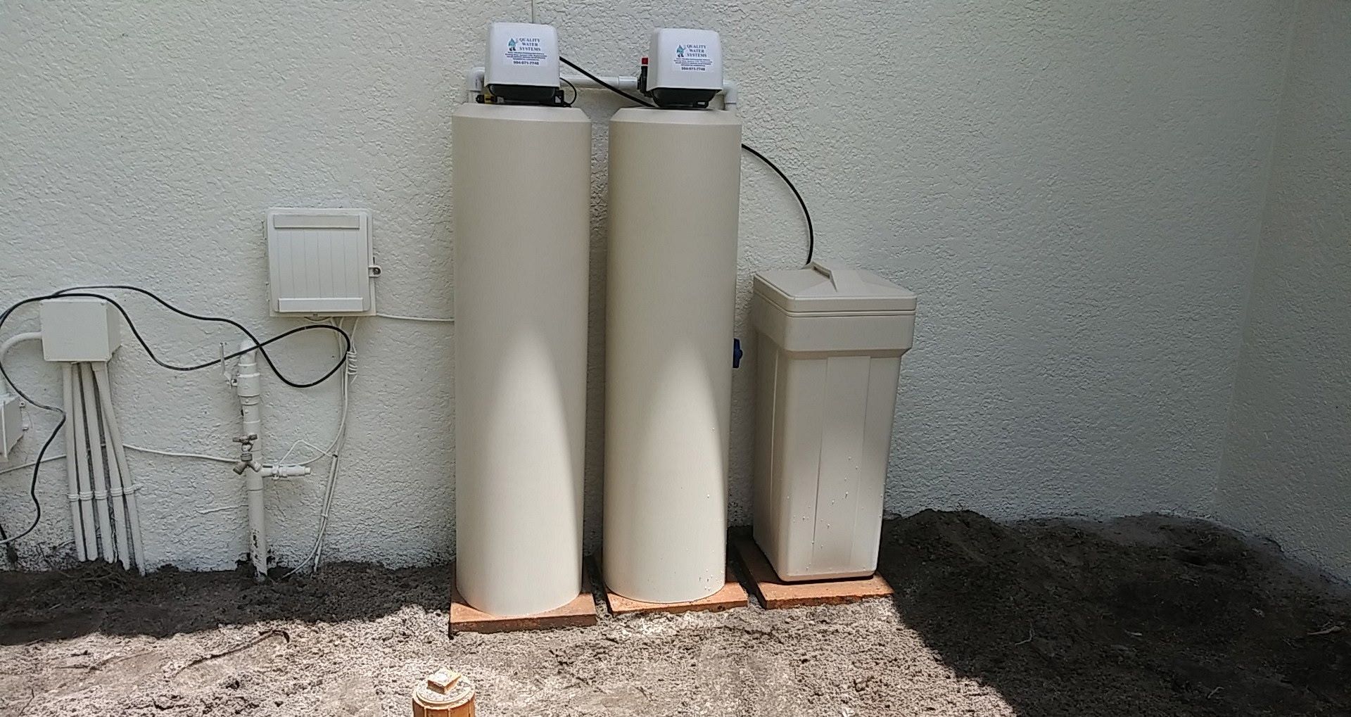 Two beige water filter tanks and a smaller unit beside a wall outdoors on gravel.