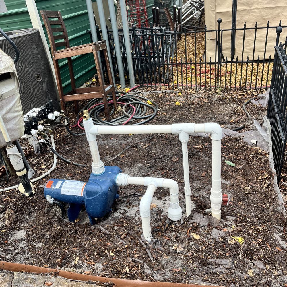 Blue water pump with white PVC pipes in a muddy yard beside a fence and garden tools.