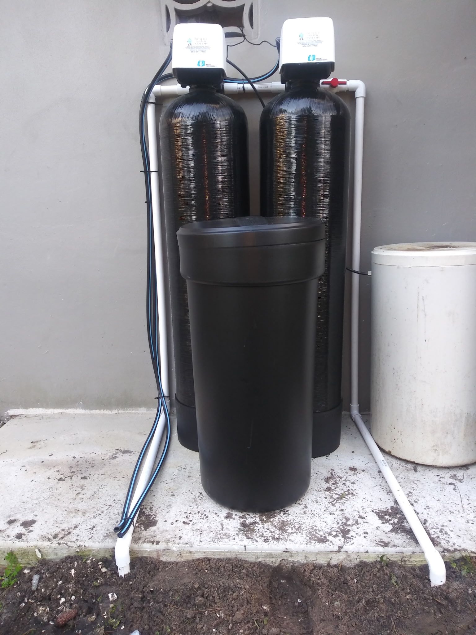 Two black water softener tanks with a black brine tank and white hoses against a gray wall.