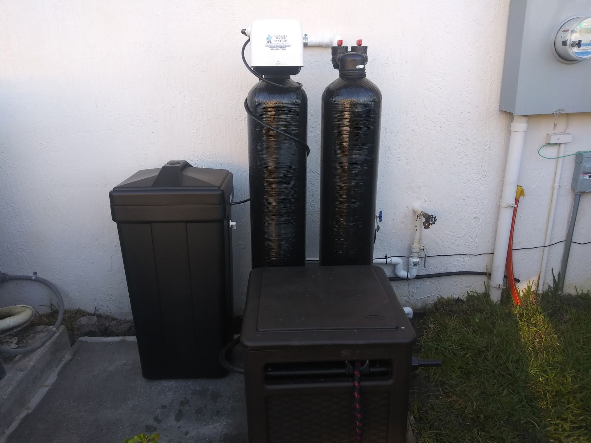 Two black water filtration tanks and a bin beside a house wall and utility box.