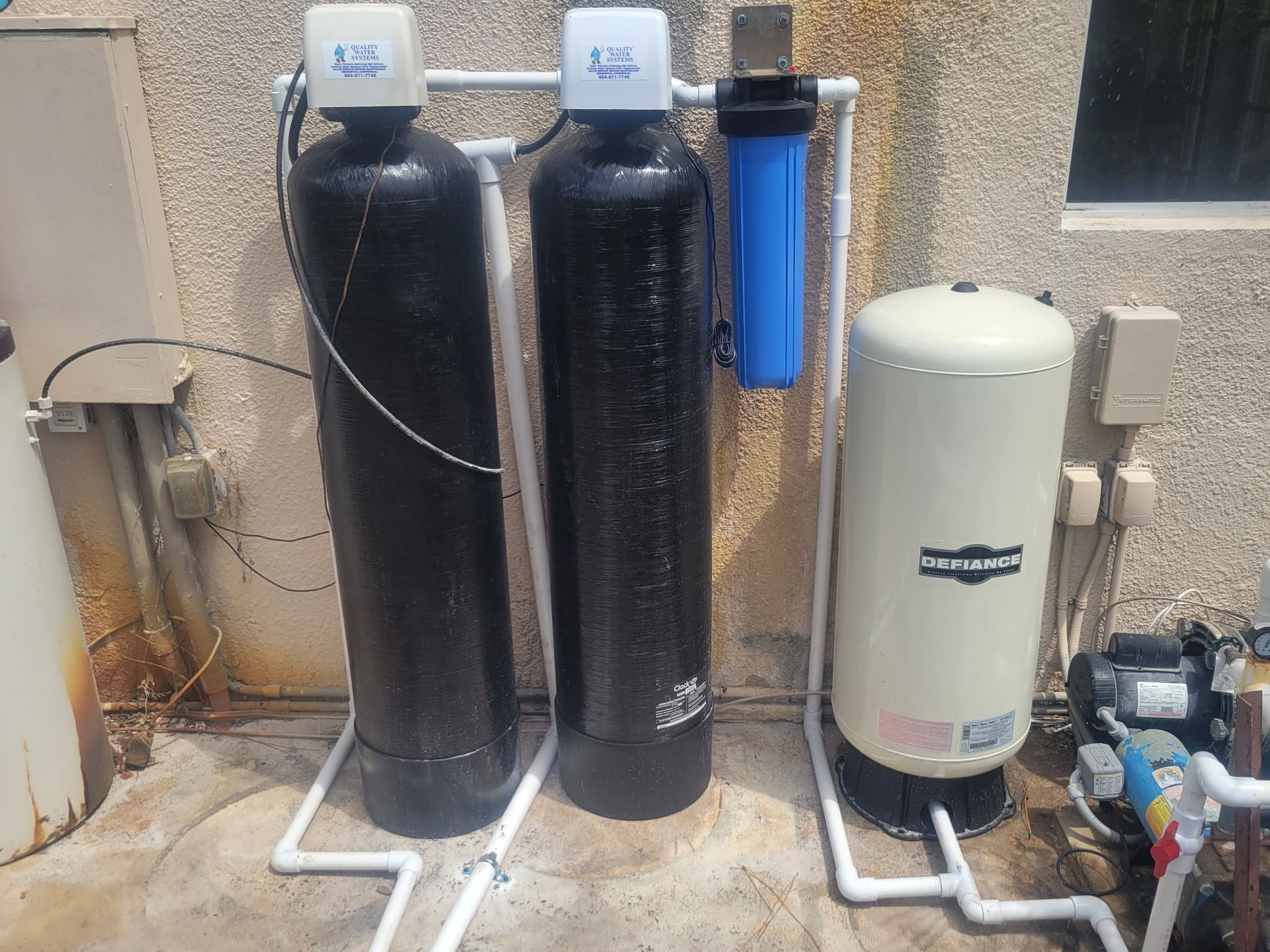 Water softener and filtration tanks beside a house exterior wall with pipes and valves