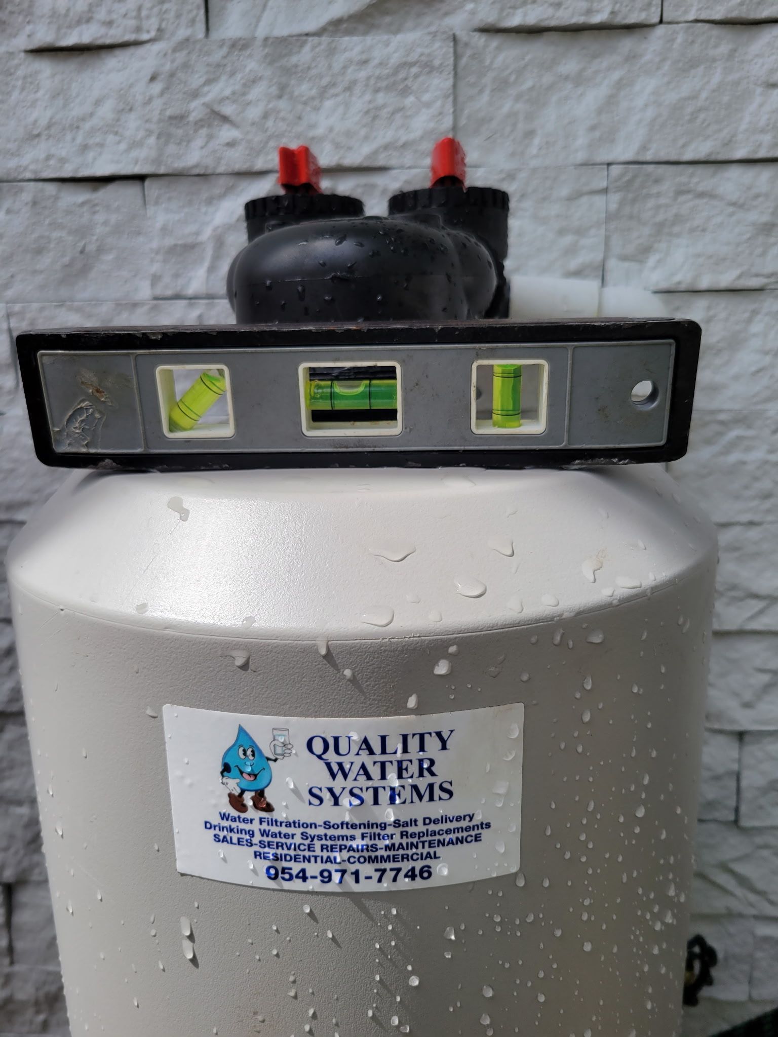 Water softener tank with control head and level indicator, branded “Quality Water Systems,” against a stone wall.