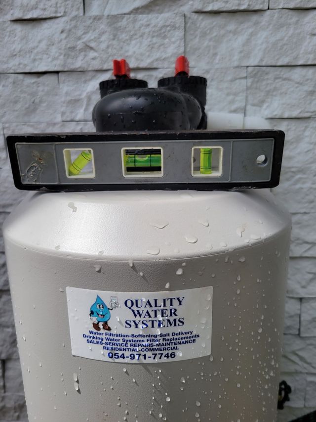 Water softener tank with control head and level indicator, branded “Quality Water Systems,” against a stone wall.
