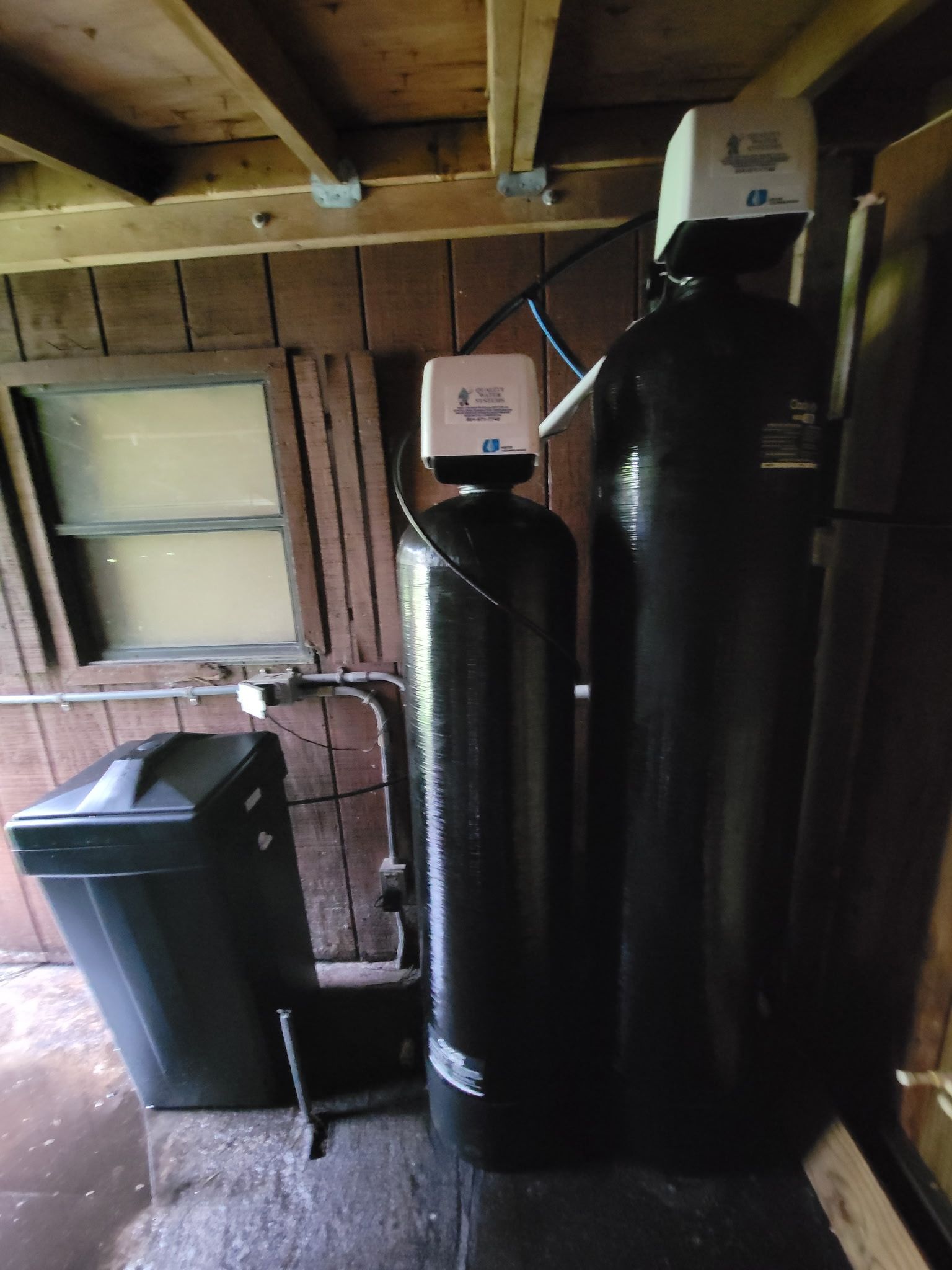 Water softener tanks and a black utility bin in a rustic wooden basement room.