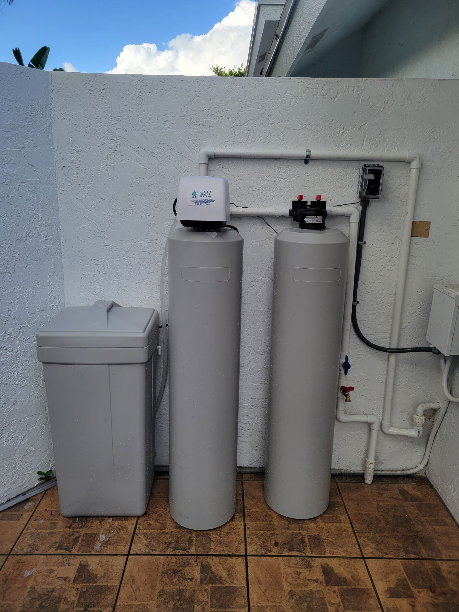 Two white water softener tanks beside a gray utility bin on a tiled patio