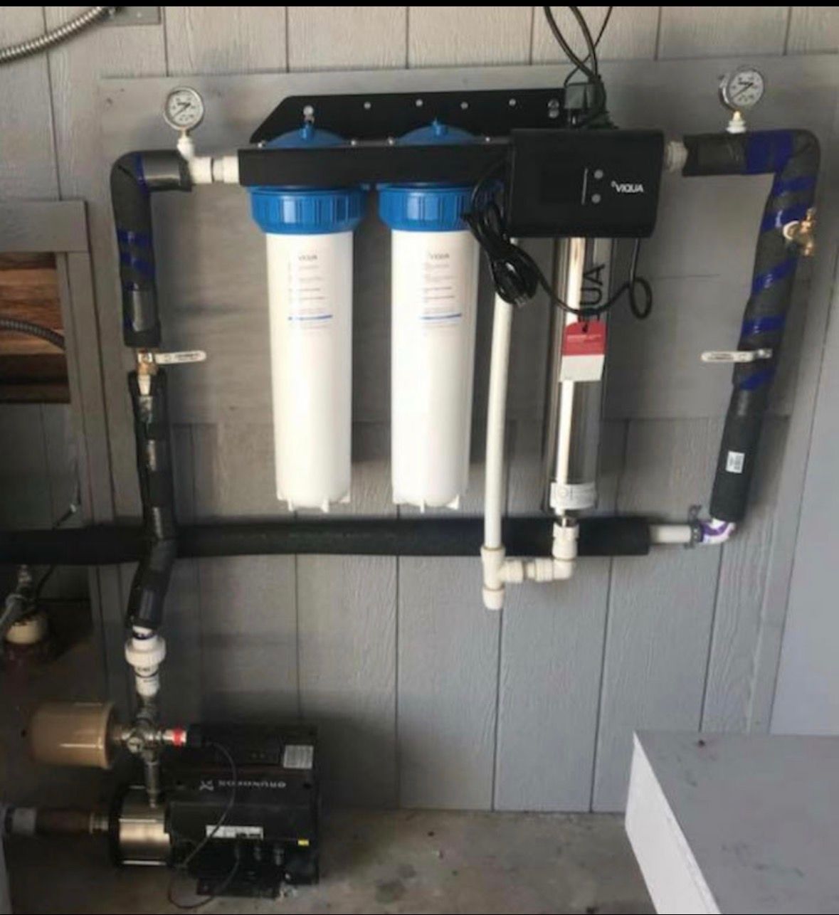 Wall-mounted water filtration system with blue and white filters, pipes, gauges, and pump in a utility room.