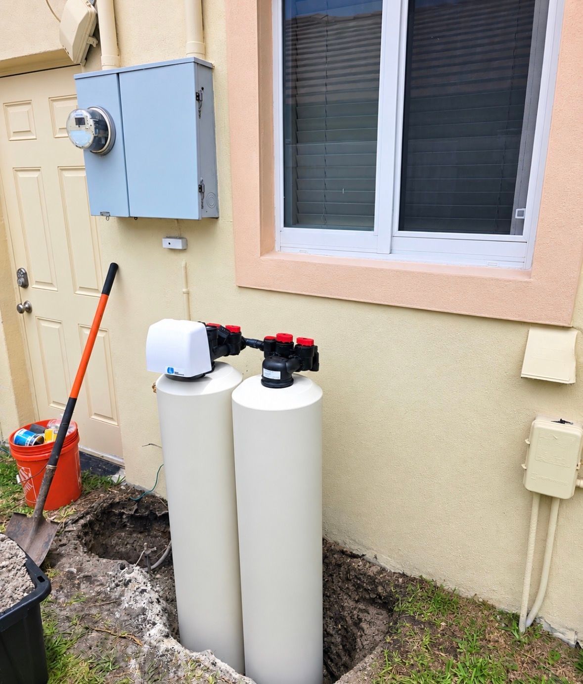 Two white water softener tanks beside a house window and electrical meter.