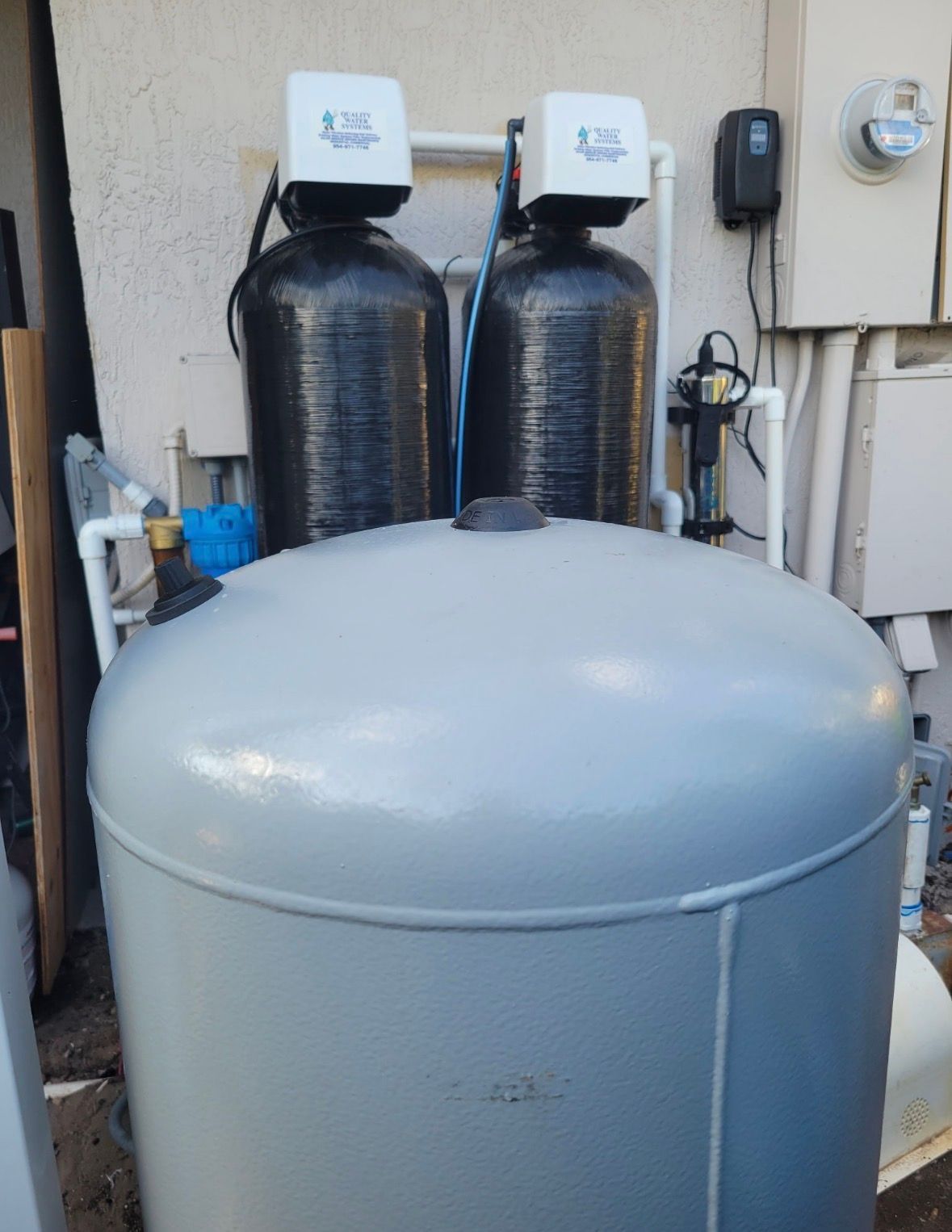 Large gray water tank in front of two black water softener tanks against a house exterior.