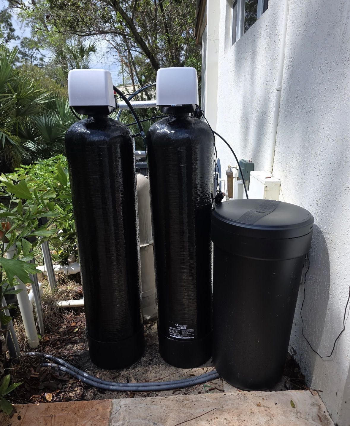 Two black water filtration tanks with white tops beside a house wall outdoors.